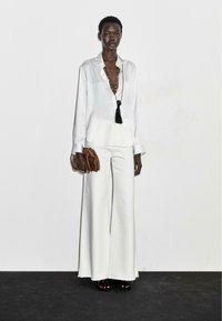 Camicia bianca in satin con bottoni, pantaloni bianchi a gamba larga e una borsa a tracolla marrone. Presenta un pendente con nappina su una lunga collana.