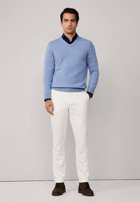 Světlomodrý pletený svetr s výstřihem do V, vrstvený přes tmavou košili s límečkem, v kombinaci s bílými kalhotami a hnědými botami. Jednoduchý design.