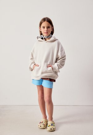 Noor tüdruk, kes kannab valget kapuutsi, siniseid lühikesi pükse, ruudulist kaelussalli ja beežikaid klotse värviliste ripatsitega, seisab ühevärvilise tausta ees.