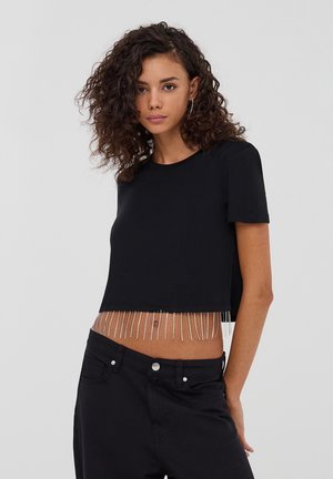 T-shirt nera cropped realizzata in tessuto liscio, con orlo a frange di perline. Maniche corte e scollatura a girocollo classica. Abbinata a pantaloni neri.