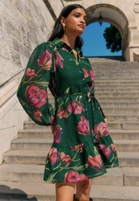 Žalia gėlėta suknelė su rožinėmis tulpėmis, mygtukų užsegimas priekyje, pūsto pynimo rankovės, diržo juosmuo ir daugiasluoksnė volanų apačia. Dėvima su auskarais.