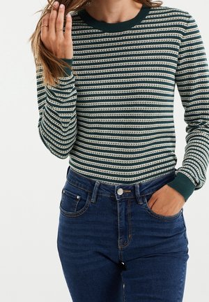 Femme portant un pull à manches longues rayé vert et blanc rentré dans un jean taille haute bleu foncé, une main dans la poche, l’autre touchant ses cheveux.