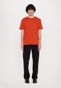Short-sleeve, crew-neck t-shirt in vibrant orange fabric, featuring a small black logo patch. Paired with black pants and shoes.