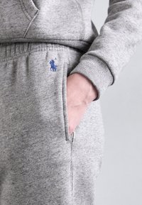 Grå bomullssweatpants med elastisk midja, med en blå broderad logga på framsidan av fickan och ribbad avslutning.