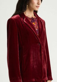 Veste en velours bordeaux avec col cranté, fermeture par un bouton unique et poches latérales, présentant une finition douce et texturée.