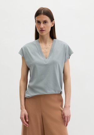 Femme aux cheveux bruns lisses portant un haut sans manches bleu clair à col en V et un pantalon taille haute beige, sur un fond blanc uni.