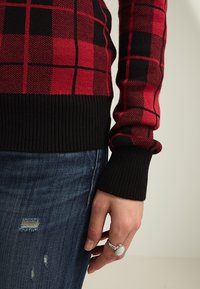 Röd och svart rutiga tröja med ribbade svarta muddar, i en texturerad stickad kvalitet. Kombinerad med slitna blå jeans.