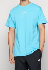 Hombre vestido con una camiseta Nike de color azul claro y pantalones Nike negros, con la mano izquierda en el bolsillo y un tatuaje en el antebrazo derecho.