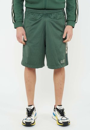 Uomo che indossa pantaloncini EA7 verde scuro e giacca con zip abbinata, in piedi con le mani rilassate lungo i fianchi, scarpe da ginnastica bianche EA7 con lacci neri.