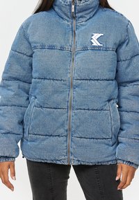 Denim puffer jacket in light blue with a high collar, zip closure, and visible stitching; features two side pockets and logo detail on chest.