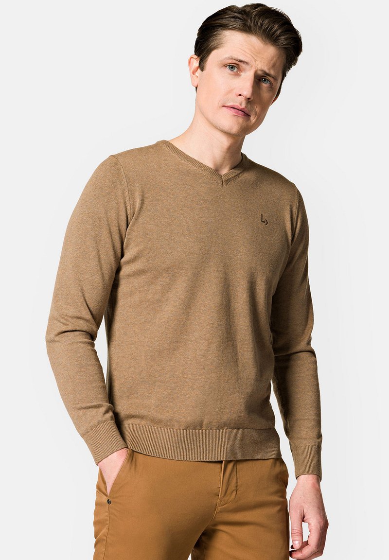 Brązowy sweter w serek z prążkowanymi mankietami i dołem, wykonany z miękkiego materiału; posiada małe logo na piersi. Noszony z brązowymi spodniami.