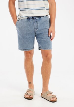 Lichtblauwe denimshorts met een trekkoord in de taille, zijzakken en afgewerkte randen, gecombineerd met beige sandalen op blote voeten.