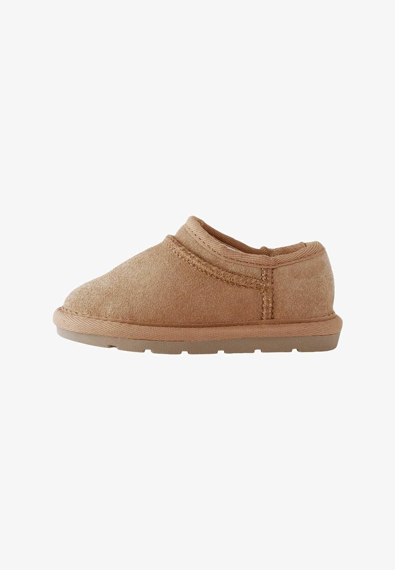 Blød brun slip-on sko med ruskindtekstur, lav hæl og syet detalje på en gummisål, vist fra siden på en hvid baggrund.
