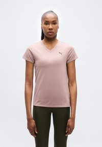 Γυναίκα με σκούρο δέρμα και μαλλιά δεμένα προς τα πίσω φοράει ένα ανοιχτό μοβ t-shirt Puma με V λαιμόκοψη και σκουρό πράσινο κολάν, στέκεται μπροστά σε άσπρο φόντο.