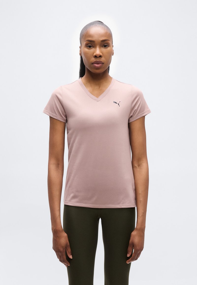 Γυναίκα με σκούρο δέρμα και μαλλιά δεμένα προς τα πίσω φοράει ένα ανοιχτό μοβ t-shirt Puma με V λαιμόκοψη και σκουρό πράσινο κολάν, στέκεται μπροστά σε άσπρο φόντο.