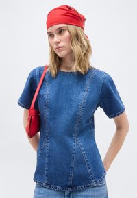 Νεαρή γυναίκα που φοράει κόκκινο μαντήλι στο κεφάλι και τζιν πουκάμισο, κρατώντας μια κόκκινη τσάντα ώμου, στέκεται μπροστά σε ένα απλό φωτεινό φόντο.