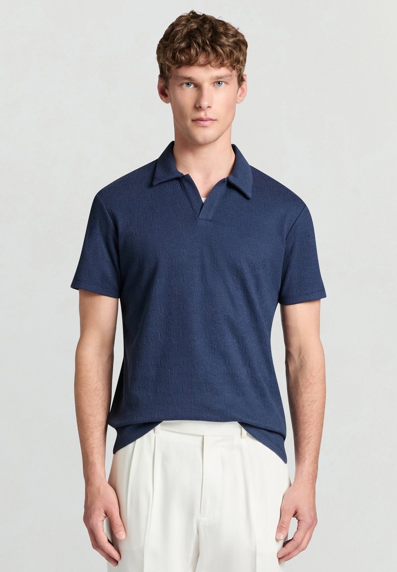 Junger Mann mit lockigem braunem Haar, der ein marineblaues kurzärmeliges Hemd mit Kragen und weiße plissierte Hosen trägt, stehend vor einem einfarbigen Hintergrund.