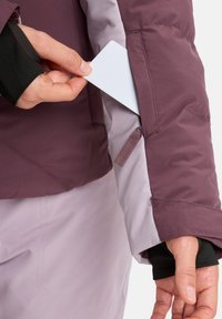 Lila und hellgraue wasserdichte Jacke mit einer Reißverschlusstasche am Ärmel, die eine Hand zeigt, die eine weiße Karte teilweise aus der Tasche zieht.