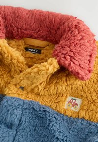 Veste en peluche colorée avec un col rouge, une section médiane jaune moutarde ornée d'un petit patch de tracteur, et une section inférieure bleue, présentant une étiquette de marque NEXT.