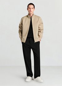 Veste bomber beige avec col montant et fermeture éclair, associée à un pantalon noir et des baskets blanches, sur un fond gris clair.