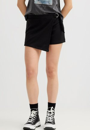 Persona que lleva una falda negra asimétrica tipo wrap con detalle de cinturón, zapatillas negras de caña alta y calcetines negros, de pie sobre un fondo blanco.