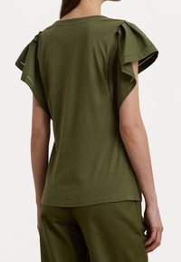 Vihreä oliivinvärinen lyhythihainen toppi, jossa on röyhelöhihat ja koristeelliset aukot reunoissa. Valmistettu pehmeästä kankaasta, löysä malli ja pyöreä pääntie.