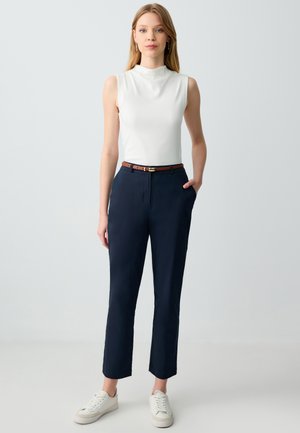 Top bianco senza maniche, pantaloni tailored blu navy con tasche laterali, cintura marrone e sneakers bianche. Tessuto liscio, forma aderente e gambe cropped.