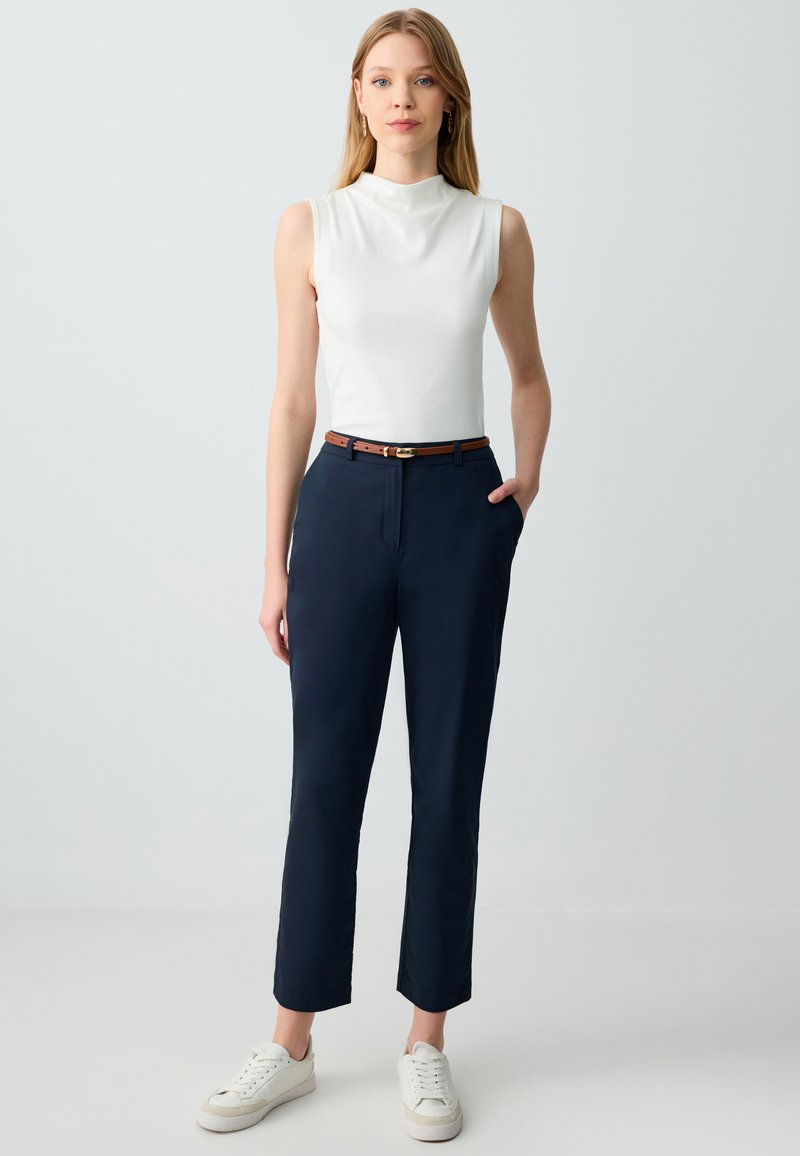 Witte mouwloze top, marineblauwe, op maat gemaakte broek met zijzakken, brown riem en witte sneakers. Gladde textuur, getailleerde pasvorm en cropped pijpen.