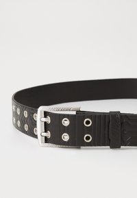 Zwarte rubberen riem met een gestructureerd ontwerp, zilveren metalen gesp met gaten en een merkaccent. Glad oppervlak met verhoogde patronen.