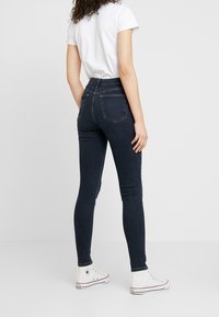 Jeans skinny de tiro alto en azul oscuro con bolsillos traseros, combinados con una camiseta blanca y zapatillas blancas, vistos desde la parte trasera.