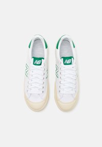 Fehér vászon sportcipők zöld részletekkel, kerek orral, fűzős záródással és texturált gumi talppal. New Balance logó az oldalain.