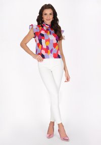 Bluză scurtă, multicoloră, cu guler înalt, având un design patterned în nuanțe de roz, roșu, galben și mov, asortată cu blugi skinny albi.