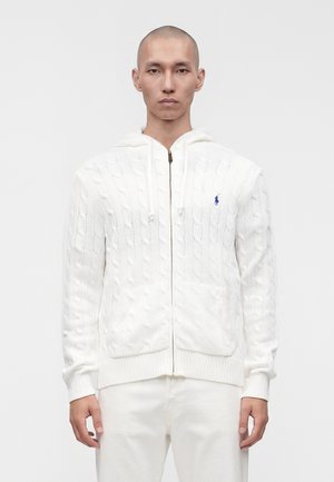 Homme portant un sweat à capuche blanc tricoté en torsades avec fermeture éclair, un petit logo brodé sur la poitrine, associé à un pantalon blanc.