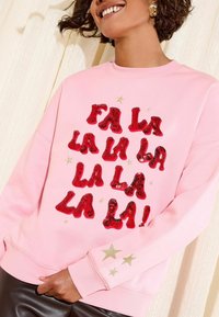Růžová mikina s červeným flitrovým nápisem "FA LA LA LA LA LA LA LA LA!" a zlatými hvězdicemi na levém rukávu. Měkčený materiál a uvolněný střih.
