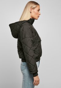 Svart quiltet jakke med hette, med diamantmønster, glidelås foran, og elastiske mansjetter. Bært over lyseblå jeans.