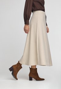 Kvinde iført en beige midikjole, brun langærmet skjorte og brune suedestøvler med blokhæl, der går på en ensfarvet baggrund.