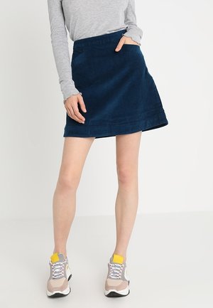 Kobieta ubrana w granatową sztruksową mini spódnicę w kształcie litery A, jasnoszary długi rękaw oraz beżowe trampki z żółtymi akcentami, stojąca na białym tle.