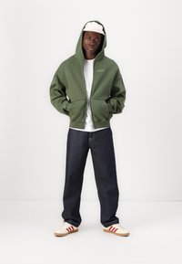 Grüne Zip-Hoodie mit Fronttaschen, getragen über einem weißen Shirt. Kombiniert mit dunkelblauen Jeans und cremefarbenen Sneakern mit roten Streifen.
