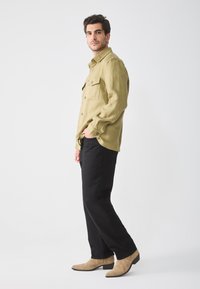 Camisa verde oliva de botones con dos bolsillos en el pecho, combinada con pantalones negros y botas de tobillo color beige. Tela suave y corte relajado.