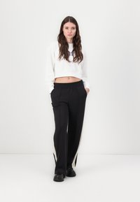 Biely skrátený sweatshirt s kruhovou grafikou a čierne nohavice s vysokým pásom, ktoré majú bočné pruhy a vrecká. Čierne topánky dotvárajú outfit.