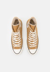 Zapatillas altas de lona color marrón claro con cordones blancos, puntera de goma y suela negra. Presentan parches de logo en los laterales.