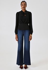 Schwarze Bluse mit langen Ärmeln und einer Schleife, kombiniert mit weit geschnittenen, dunkelblauen Jeans. Das Outfit hat eine taillierte Passform und eine glatte Stofftextur.