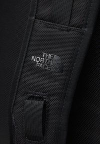 Gros plan d'une sangle de sac à dos noire texturée avec le logo The North Face brodé en fil blanc.