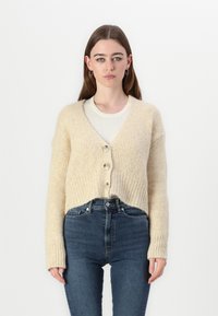 Cardigan en tricot beige avec un col en V, trois grands boutons, texture côtelée et manches longues, associé à un jean bleu taille haute.