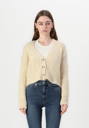 Cardigan en tricot beige avec un col en V, trois grands boutons, texture côtelée et manches longues, associé à un jean bleu taille haute.