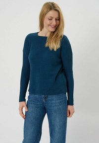 Türkisfarbenes Rippstrickpullover mit langen Ärmeln, rundem Ausschnitt und lässiger Passform, kombiniert mit hellblauen gerade geschnittenen Jeans.