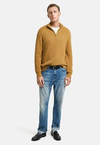 Maglione a maglia color senape con un motivo testurizzato, con zip a un quarto, abbinato a jeans azzurri chiari e mocassini neri. Vestibilità casual, accenti di design minimi.