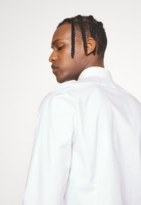 Chemise blanche à bouton avec un col structuré et un tissu lisse, portée par une personne de dos, montrant des cheveux tressés et un design minimaliste.