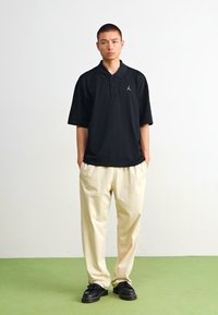 Čierny polo s krátkym rukávom a šedým logom, kombinovaný s svetlými béžovými nohavicami a čiernymi topánkami. Jednoduchý dizajn, voľný strih a hladká textúra.