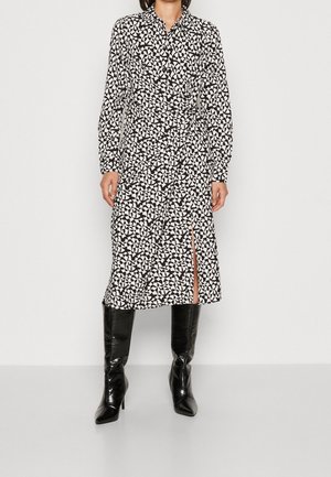Woman wearing a long-sleeve black and white patterned midi dress with a side slit, paired with black shiny heeled knee-high boots.